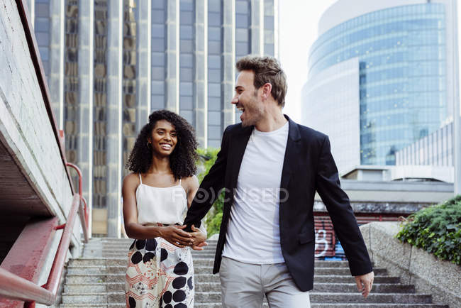 Handsome man smiling and looking at lovely African-American woman while walking down stairs together on city street — Stock Photo