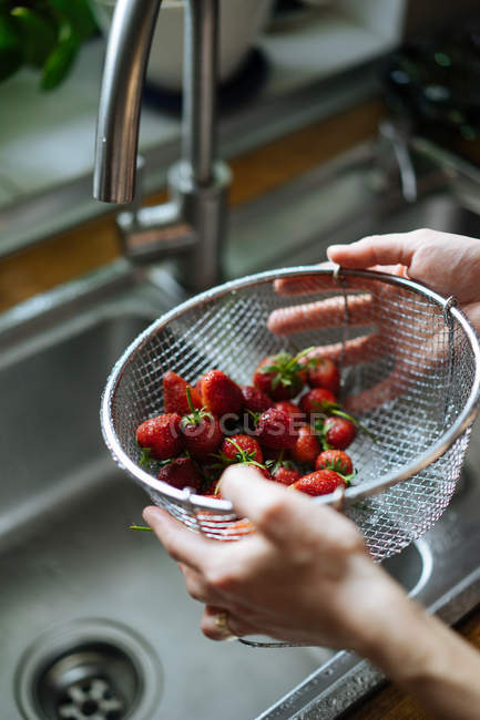 Mãos humanas segurando filtro de morangos frescos sob torneira pia na cozinha — Fotografia de Stock