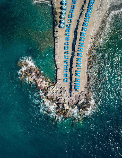 Вид с воздуха на скалистый и песчаный полуостров с ярко-голубыми зонтиками и пляжными креслами в окружении прекрасного океана — стоковое фото
