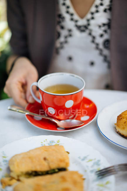 Main féminine tenant une tasse de thé en céramique rouge à pois sur une soucoupe sur une table de jardin — Photo de stock