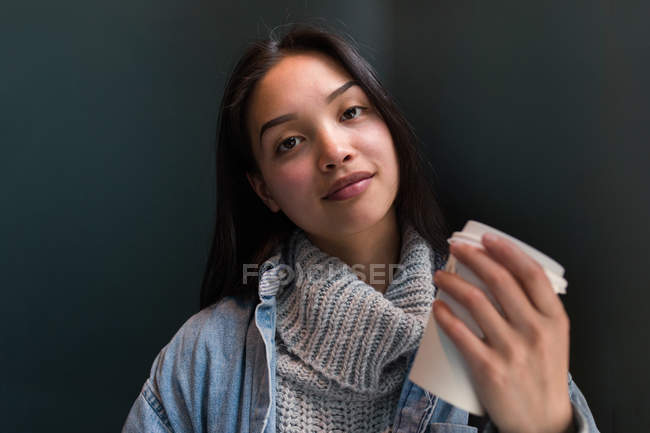 Mujer étnica bonita en suéter y chaqueta de mezclilla sosteniendo taza de papel de café y mirando a la cámara - foto de stock