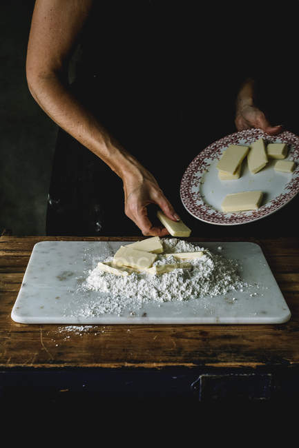 Cropped shot of woman adding butter pieces to pile of flour on board while making dough — Stock Photo