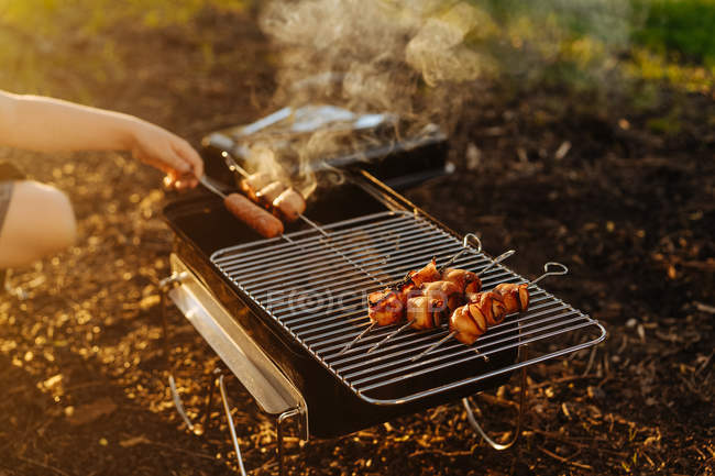 Mão humana preparando bacon e salsichas em espetos grelhando na queima de carvão em grelha portátil ao ar livre — Fotografia de Stock