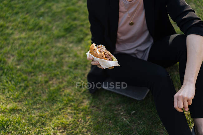 Mujer sentada en la hierba en el parque y comiendo hamburguesa para llevar - foto de stock