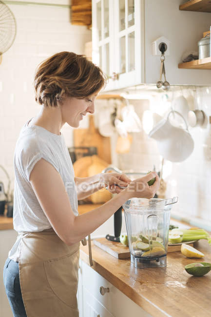 Mujer poniendo ingredientes en la taza de plástico de la licuadora para hacer batido verde saludable - foto de stock