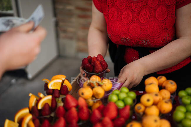 Vista de la cosecha de la hembra poniendo taza de fresas jugosas maduras en un paquete en el mostrador con frutas frescas brillantes y bayas con la mano sosteniendo el dinero cerca - foto de stock