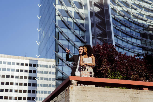 Multiracial couple hugging and pointing in front of modern buildings — Stock Photo