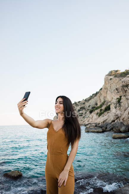 Mujer elegante tomando selfie en la costa rocosa por el agua de mar - foto de stock