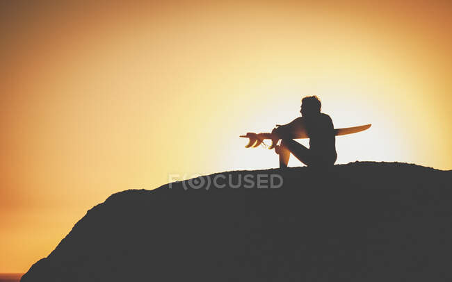 Vista lateral del surfista masculino figura oscura sentada en la colina y sosteniendo la tabla en las manos en el fondo iluminado por la espalda al atardecer - foto de stock