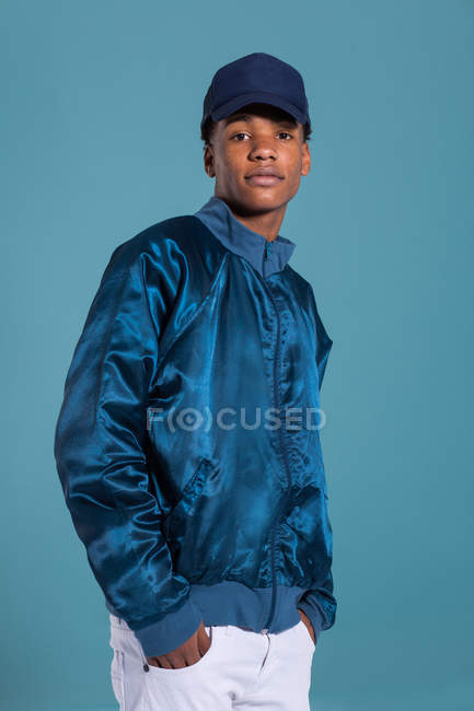 Trendy ethnic man wearing shiny blue bomber jacket and cap against blue background — Stock Photo