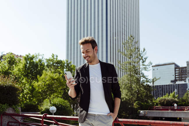 Elegant guy using smartphone while leaning on railing on street of modern city — Stock Photo