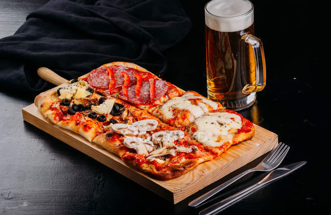 Pizza cortada con queso, salsa y berenjenas en rodajas sobre tabla de madera sobre mesa oscura con vaso de cerveza - foto de stock