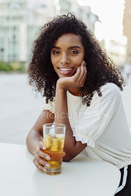 Charming African-American woman holding glass of beverage in outdoor cafe — Stock Photo