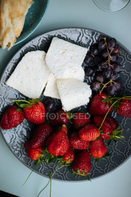 Гарний набір свіжого м'якого сиру з полуницею та вишнями, що лежать на текстурованій темній тарілці на синьому столі зверху — стокове фото