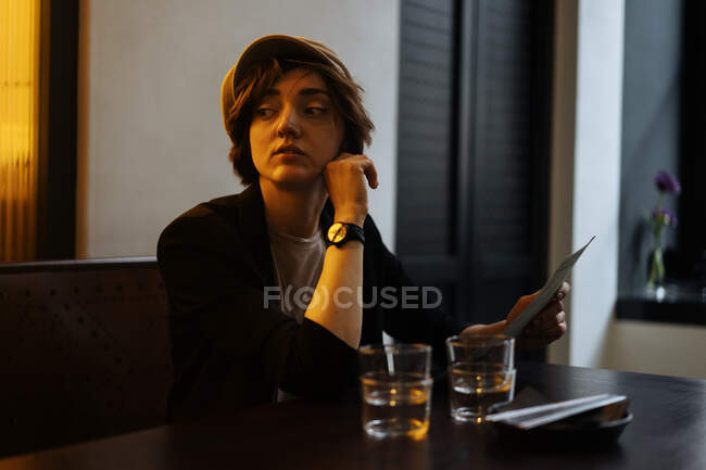 Молода красива жінка в кепці, сидячи за столом в сучасному ресторані і читаючи через меню — стокове фото