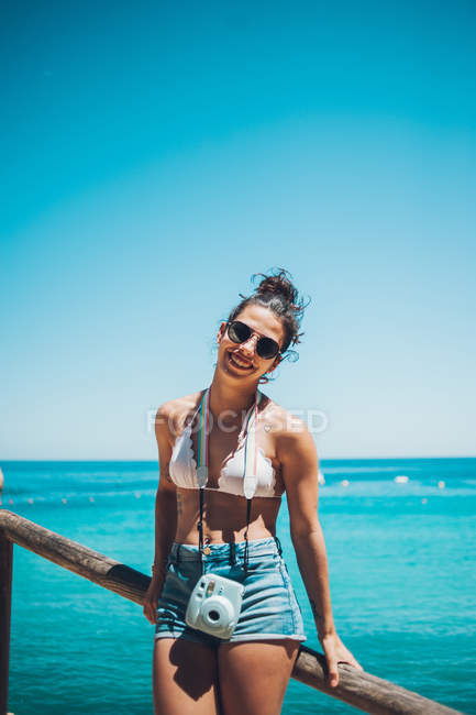 Sonriente joven con ropa de verano apoyada en pasamanos de madera en la playa y mirando a la cámara - foto de stock