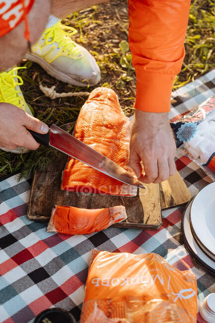 Crop shot from above of man cutting and slicing salmon fillet on board on the ground for picnic meal in sunlight — Stock Photo