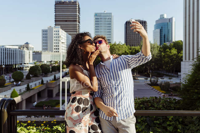Весела багаторасова пара позує за селфі, стоячи на тлі сучасного міста — стокове фото