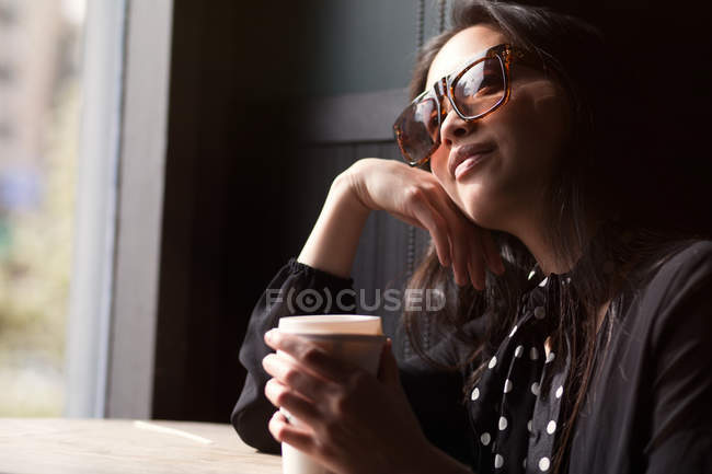 Mujer sonriente elegante de moda en gafas de sol y pañuelo sosteniendo taza de papel de café y apoyándose en la mesa - foto de stock