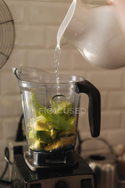 Verser de l'eau du pichet en céramique dans une tasse en plastique de mélangeur remplie de mélange de fruits et légumes pour smoothie — Photo de stock