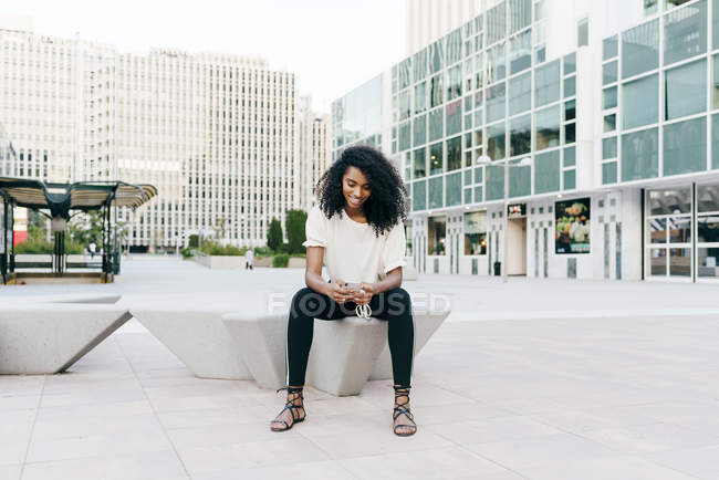 Улыбающаяся афроамериканка, сидящая на городской улице и держащая смартфон — стоковое фото
