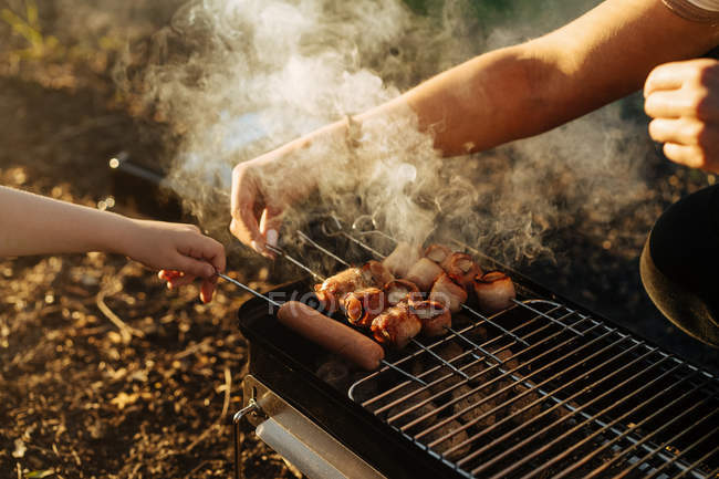Mãos humanas preparando bacon e salsichas em espetos grelhando em carvão em chamas em grelha portátil ao ar livre — Fotografia de Stock