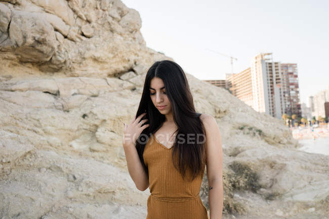 Beautiful stylish brunette touching hair and looking down on rocky coast — Stock Photo