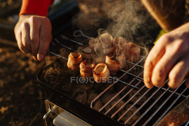 Mãos masculinas preparando bacon e salsichas em espetos grelhando na queima de carvão em grelha portátil ao ar livre — Fotografia de Stock