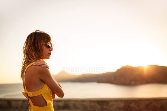 Жінка в жовтій сукні та сонцезахисних окулярах дивиться на узбережжя під час заходу сонця — стокове фото