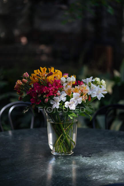 Eleganti fiori variopinti assortiti in bouquet in piedi in vaso di vetro con acqua su tavolo rotondo nero illuminato dal sole con piante su sfondo sfocato — Foto stock
