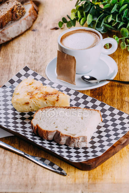 Coffee in white porcelain cup on saucer with checkered chopping board with slice of bread and piece of pastry — Stock Photo
