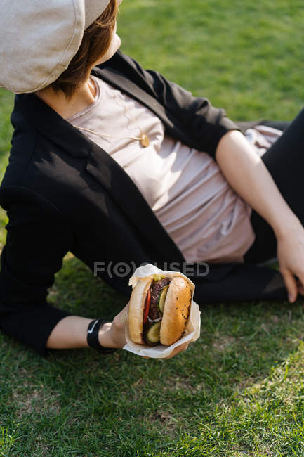 Elegante donna rilassante su erba nel parco e tenendo hamburger da asporto — Foto stock