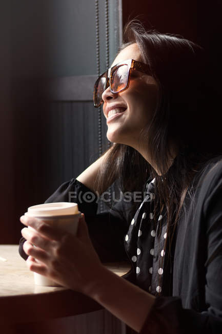 Mujer sonriente elegante de moda en gafas de sol y pañuelo sosteniendo taza de papel de café y apoyándose en la mesa - foto de stock