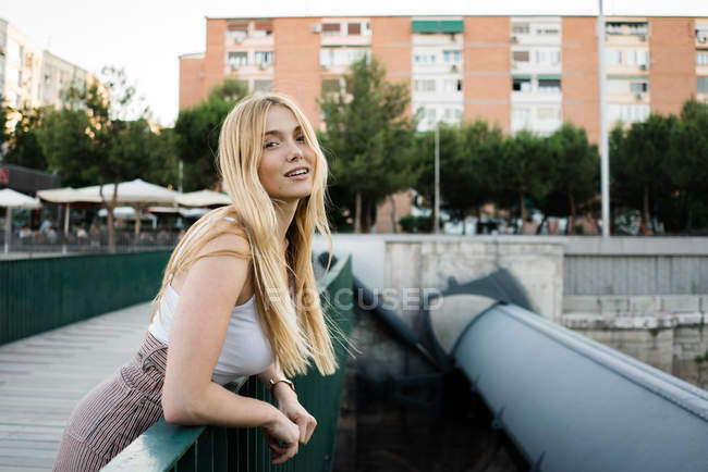 Porträt einer attraktiven jungen Frau, die lächelt und in die Kamera blickt, während sie sich an das grüne Geländer der Brücke über den Stadtfluss lehnt — Stockfoto