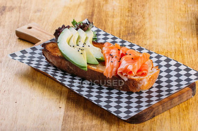 Fisch- und Avocado-Sandwich auf kariertem Brett auf Holztisch — Stockfoto