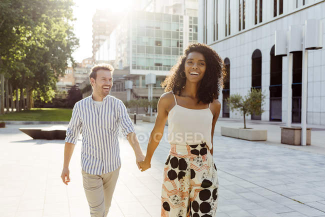 Sonriente pareja multirracial caminando de la mano en la calle de la ciudad en un día soleado - foto de stock