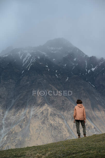 Вид ззаду чоловіка в теплому одязі з рюкзаком пішохідний в горах, що стоять на траві, дивлячись на зубчастий гірський хребет, вкритий снігом і вершинами, схованими в хмарах — стокове фото
