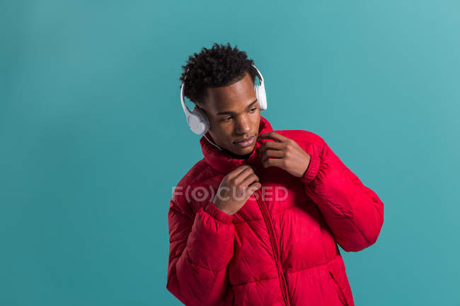 Elegante hombre afroamericano con auriculares y chaqueta hinchada roja de pie sobre fondo azul - foto de stock