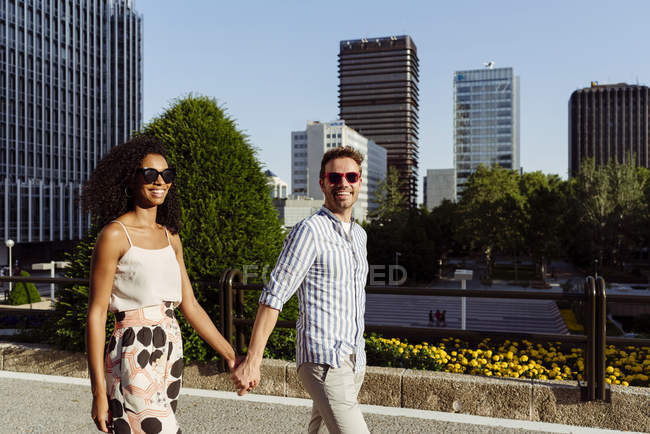Счастливая многорасовая пара, улыбающаяся и держащаяся за руки во время прогулки по городской улице вместе в солнечный день — стоковое фото