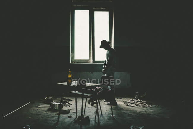 Silueta de hombre en sombrero de pie cerca de las ventanas en la habitación sucia con basura
. - foto de stock