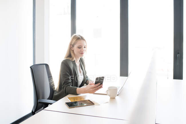 Jovem mulher loira elegante sentada à mesa com laptop e xícara de café usando smartphone à luz do dia — Fotografia de Stock