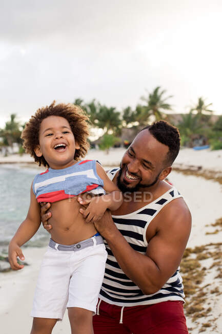 Happy African-American man climbing up little son while having fun on sandy beach together — Stock Photo