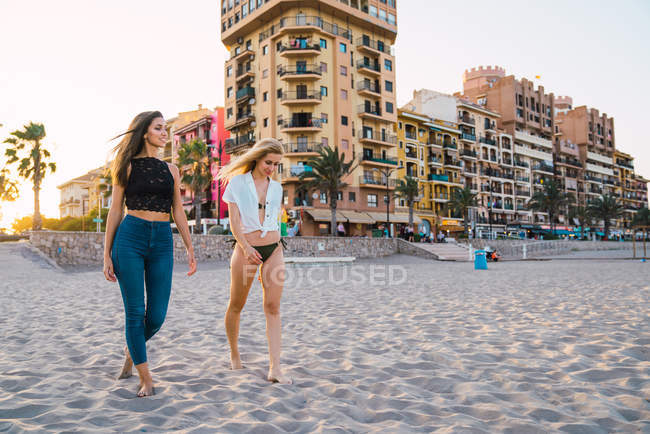 Улыбающиеся подруги прогуливаются по пляжу со зданиями на заднем плане — стоковое фото