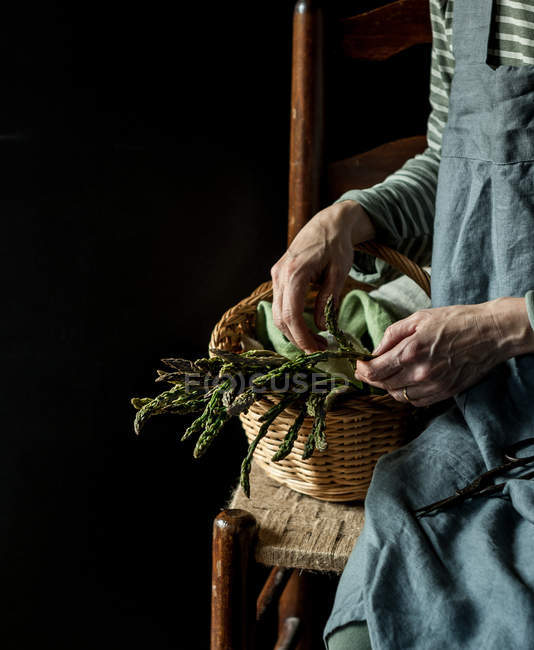 Human hand taking asparagus from basket on chair on black background — Stock Photo