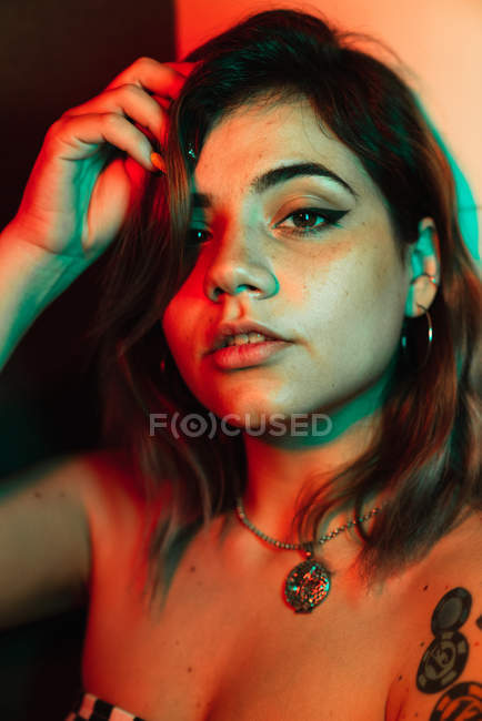 Молодая женщина трогает волосы и смотрит в камеру, стоя в комнате, освещенной красным и зеленым светом во время вечеринки — стоковое фото