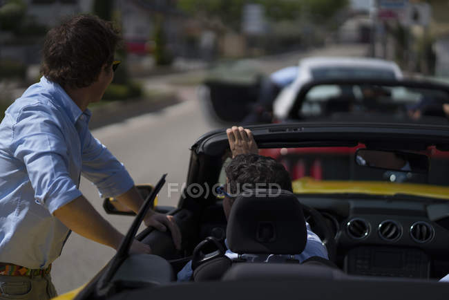 Hombre sentado en un coche nuevo moderno y hablando con el hombre de pie cerca y apoyado en el coche - foto de stock
