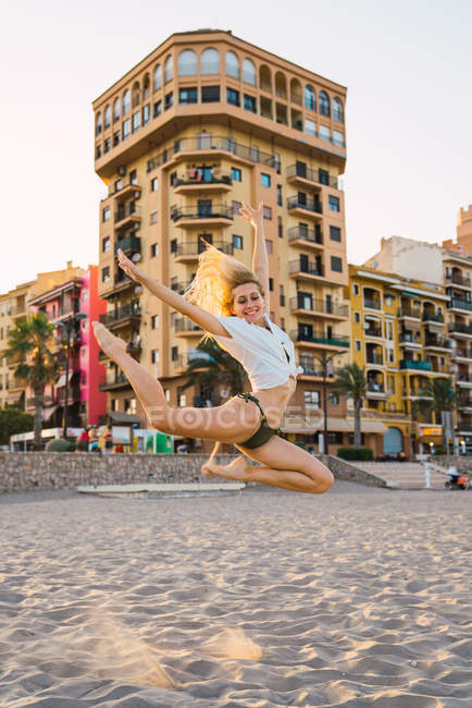 Гнучка молода жінка стрибає на пляжі з будівлями на фоні — стокове фото