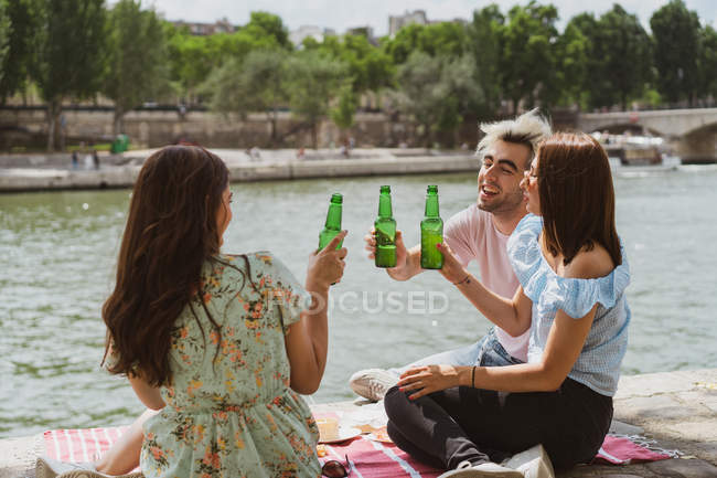 Smiling male and female friends relaxing on waterfront and clinking with bottles — Stock Photo