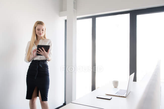 Mujer elegante que usa traje y tableta mientras está de pie en la luz del día brillante dentro de la oficina moderna - foto de stock
