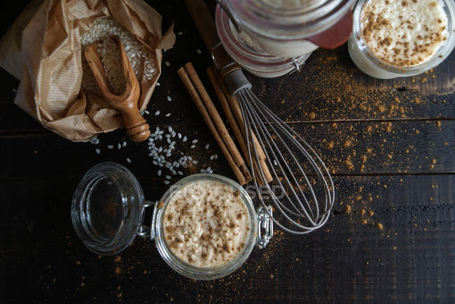 Стеклянные банки сладкого десерта риса с молоком и корицей на деревянном столе с ингредиентами — стоковое фото